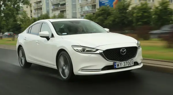 Biała Mazda 6 2.0 benzyna jedzie po mokrej drodze, w tle budynki i drzewa.