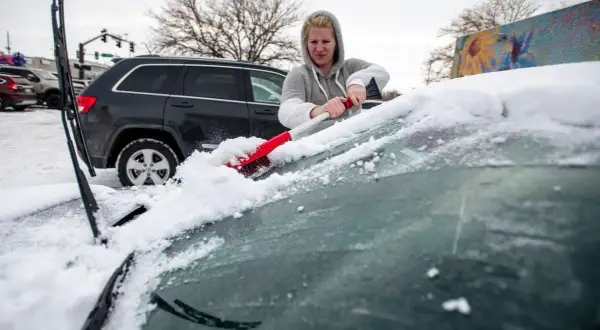 Kvinna skrapar snö från bilrutan med en röd borste. Snön ligger tjock på bilens framruta.