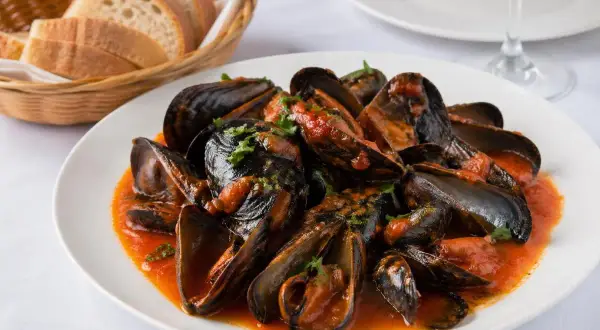 Mejillones a la gallega servidos en un plato blanco con salsa de tomate y perejil picado. Al fondo, una cesta de pan y una copa de vino.