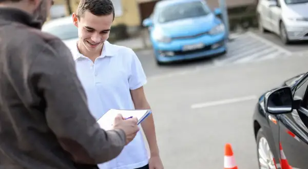 Młody mężczyzna z aparatem na zębach uśmiecha się, gdy instruktor jazdy zapisuje coś na kartce. Niedługo dowie się, od ilu lat można jeździć autem.