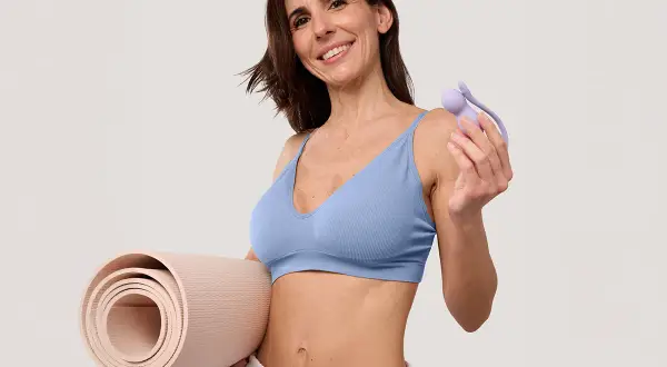 Une femme souriante tient un tapis de yoga et un accessoire pour la musculation du périnée, suggérant quels sont les sports favorables à la musculation du périnée.