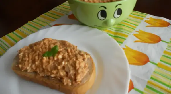 Pyszna pasta z makreli i twarogu na kromce chleba, ozdobiona listkiem bazylii. Obok miseczka z pastą i wesoła buźka.