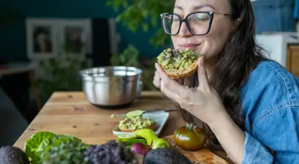 Frau genießt Avocado-Toast, ein gesunder Snack für das avocado abnehmen. Frische Zutaten liegen auf dem Tisch.