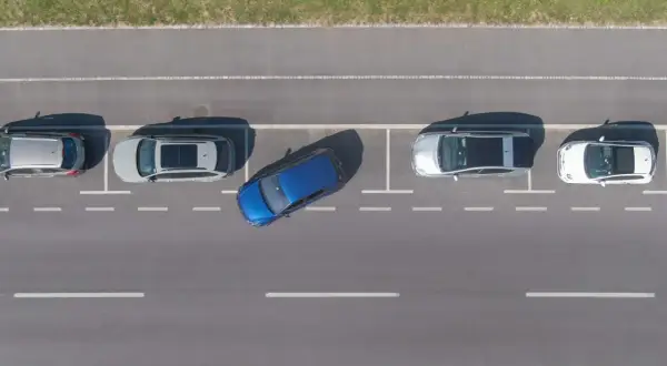 Niebieski samochód wykonuje parkowanie równoległe tyłem między dwoma innymi autami na parkingu.