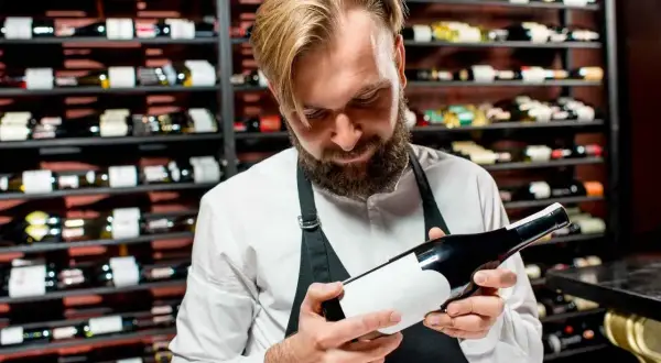 Un sommelier esamina una bottiglia, un passo fondamentale per capire se un vino è buono. Dietro di lui, scaffali pieni di bottiglie.