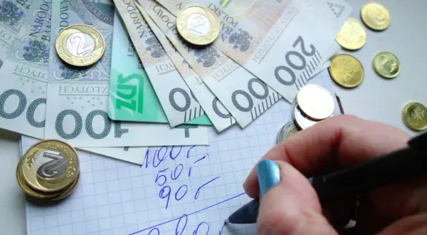 Ręka z niebieskim lakierem pisze na kratkowanym papierze, obok leżą polskie banknoty i monety. Planowanie wydatków to pierwszy krok do rewaloryzacji budżetu.