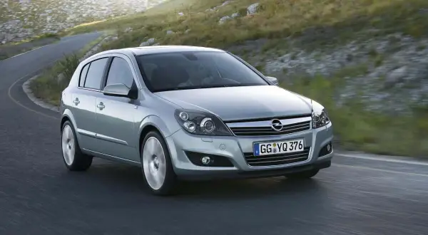Srebrny Opel Astra 2005 na krętej drodze, otoczony zielonymi wzgórzami.
