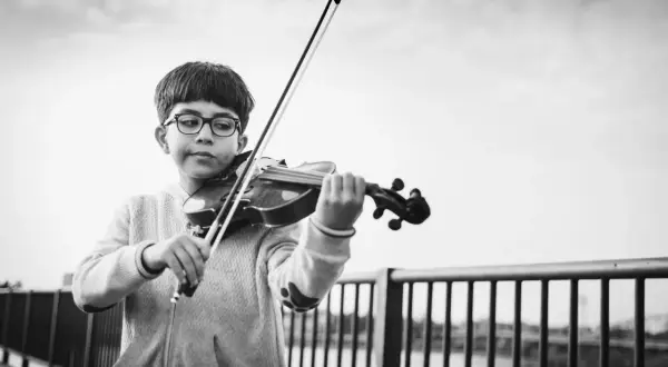 Ein Junge mit Brille spielt konzentriert Geige. Er steht an einer Brüstung, die Musik erklingt im Freien.