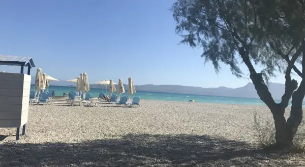 Słoneczny dzień na plaży Alkyon. Białe leżaki i parasole czekają na plażowiczów. W tle turkusowe morze i góry.
