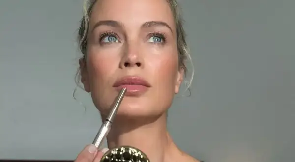 Kobieta z niebieskimi oczami i blond włosami nakłada błyszczyk. Jej skóra promienieje naturalnym blaskiem, idealnym do makijażu glow.