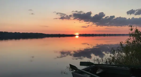 Łódź czeka nad spokojną rzeką białych nocy, odbijając pomarańczowe niebo i chmury.