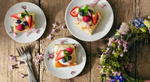 Tre tallrikar med vegansk midsommarmat, toppad med bär och blommor, ligger på ett träbord. En blomsterkrans ligger bredvid.