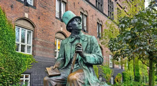 Pomnik Hansa Christiana Andersena w Kopenhadze, siedzącego z książką i laską, na tle ceglanego budynku porośniętego bluszczem.
