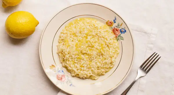 Un cremoso risotto al limon, con ralladura de limón espolvoreada, servido en un plato floral. Dos limones frescos reposan cerca.