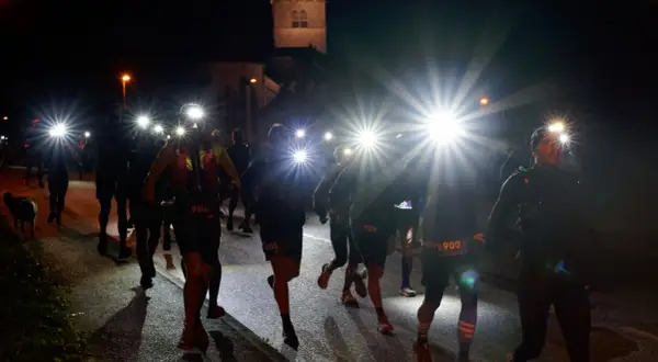 Courseurs de trail de nuit, lampes frontales allumées, traversant un village sombre. Un chien les accompagne.