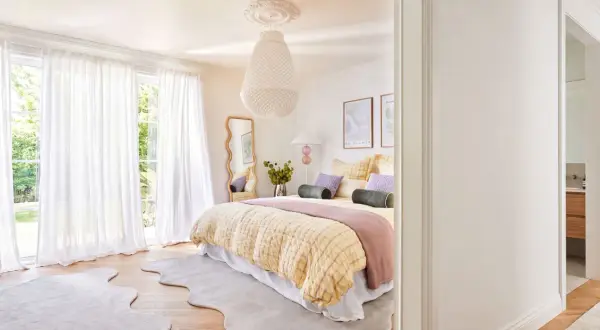 Ein geschwungener Spiegel im Schlafzimmer reflektiert das helle Zimmer mit Bett, weißen Vorhängen und Blick ins Grüne.