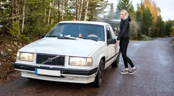 Vit Volvo med en tjej som öppnar dörren. En svensk registreringsskylt syns tydligt på bilen.