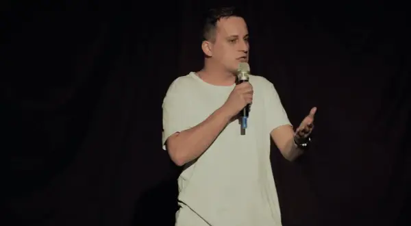 Mężczyzna w białej koszulce stoi na scenie z mikrofonem w dłoni, gestykulując.