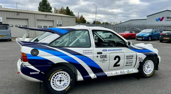 En vit rallybil Ford Escort Cosworth med blåa detaljer och nummer 2. Bilen står parkerad utomhus.