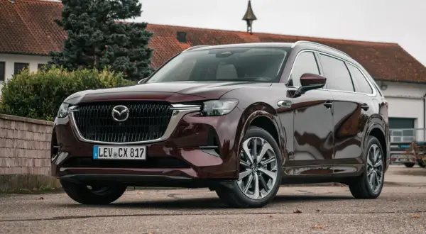 Elegancka, ciemnobordowa Mazda CX-80 stoi przed budynkiem z czerwoną dachówką.