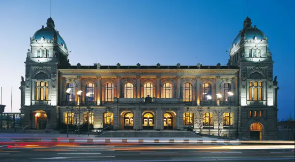 Prachtvolle alte Stadthalle im Abendlicht, beleuchtete Fenster und Türme, Lichterspuren auf der Straße.