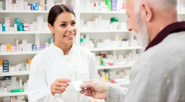 Uśmiechnięta farmaceutka podaje starszemu panu opakowanie leków. W tle apteczne półki pełne medykamentów, tworzące poltixa.