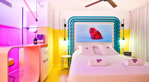 Pokój w stylu memphis z łóżkiem, lodówką Smeg i obrazem czerwonego kota. Kolorowe ściany i meble tworzą radosną atmosferę.