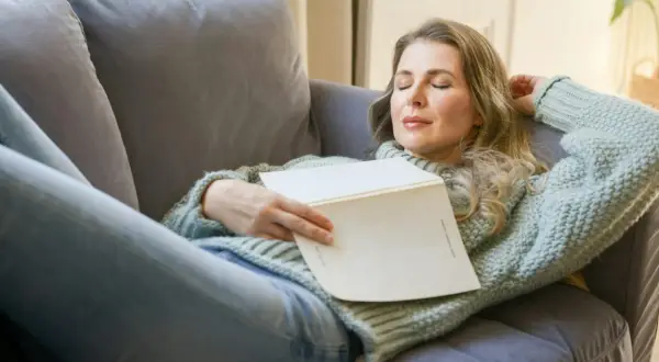 Kvinna vilar på soffan med en bok i famnen, drömmer om hur lång ska en powernap vara för maximal återhämtning.