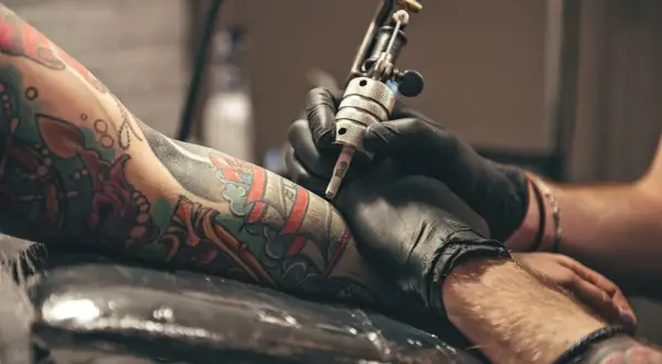 Manos enguantadas trabajan en un tatuaje colorido en un brazo. El artista usa una máquina de tatuar, creando arte permanente.