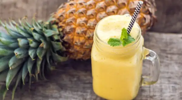 Orzeźwiający smoothie z ananasem, ozdobione miętą, obok świeżego owocu na drewnianym tle.