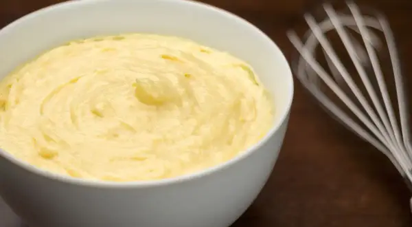 Crema pasticcera senza glutine, liscia e vellutata, pronta per farcire dolci. Vicino, una frusta in movimento.