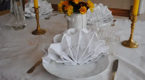 Vackert dukat bord med vita, vikta servetter i glas. Guldljusstakar och blommor skapar en festlig stämning.