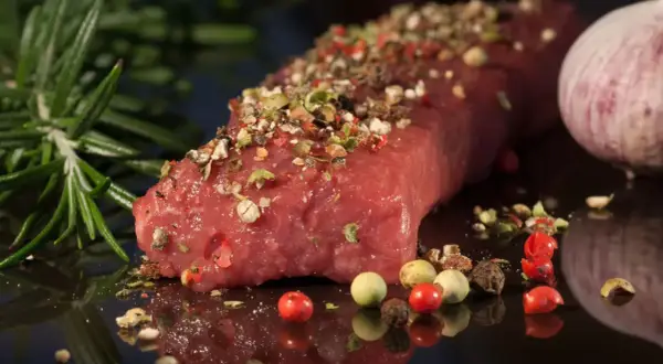 Un pezzo di carne succulenta, condita con pepe misto e rosmarino, pronta per diventare una deliziosa carne tipica pugliese. Accanto, uno spicchio d'aglio.