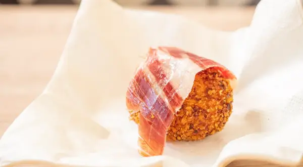 Croquetas de jamón crujientes, envueltas en una loncha de jamón serrano, servidas sobre papel blanco.