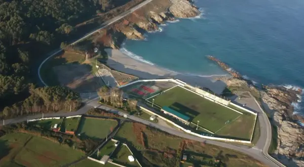 Vista aérea de un campo de fútbol junto a una playa a marosa, con el mar azul y rocas.