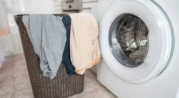 Cesto della biancheria con capi grigi e beige accanto a una lavatrice. Come risolvere quando un capo perde colore?