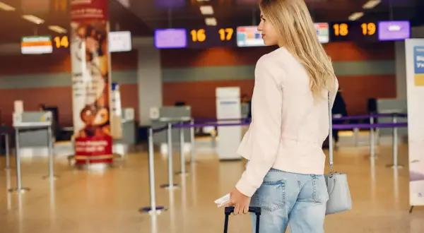Kobieta z walizką i biletami czeka na odprawę na lotnisku w Katowicach. W tle widać tablice z numerami stanowisk.