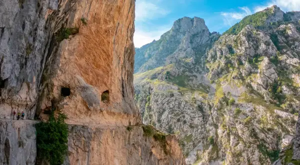 Senderos tallados en roca, con personas caminando. El inicio y fin de la ruta del Cares se pierden entre imponentes montañas verdes y cielos azules.