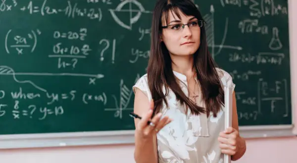 Nauczycielka matematyki w okularach, wyjaśniająca wzory na tablicy. Dofinansowanie do okularów dla nauczycieli to ważna sprawa.
