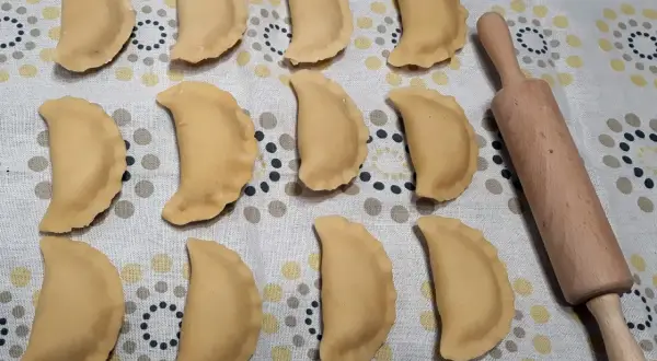 Gotowe do gotowania pierogi z jabłkami, ułożone rzędami na wzorzystym obrusie, obok drewnianego wałka.