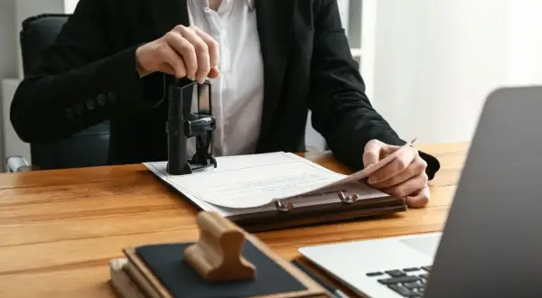 Osoba w garniturze stempluje dokumenty, być może związane z hipoteką. Na biurku leżą też książki i laptop.