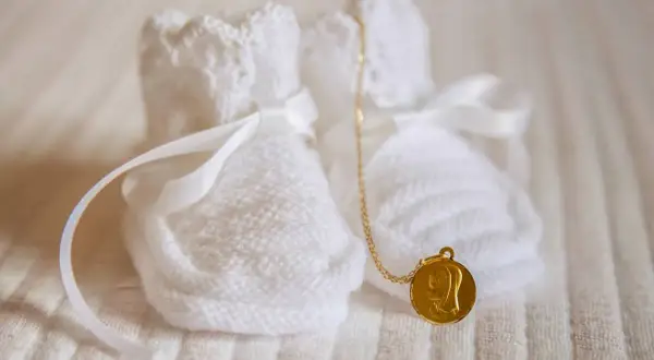 Douces chaussons blancs tricotés, ornés de rubans, avec une médaille dorée de la Vierge. Un symbole de marraine sans baptême, plein d'amour.