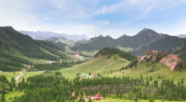 Grüne Hügel, Nadelwälder und Felsformationen prägen diese weite mongolische Landschaft. Kleine Gehöfte und Jurten sind in der Ferne zu erkennen.