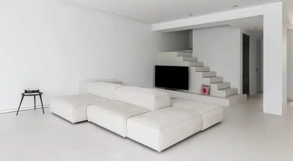 Soggiorno minimalista con divano modulare bianco e pavimento in resina epossidica opaca. Scale moderne conducono a un'area con TV.