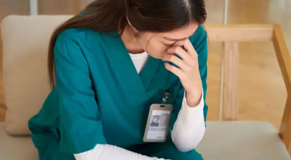 Une infirmière en blouse verte, le visage caché dans ses mains, semble lutter pour trouver son équilibre émotionnel.