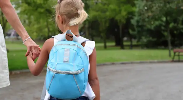 Dziecko w niebieskim plecaku trzyma za rękę dorosłego, idąc przez park.