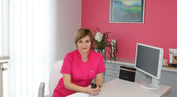 Lekarka w różowym uniformie siedzi przy biurku w gabinecie dermatologicznym.