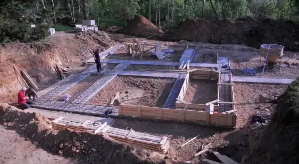 Budowa fundamentów domu. W wykopie widać zbrojenie i szalunki. Pracownicy przygotowują teren pod budowę, a chudziak czeka na beton.
