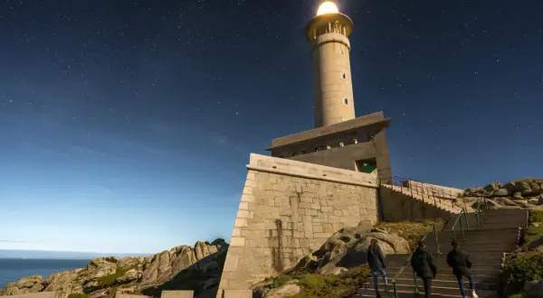 El faro de Punta Nariga ilumina la noche estrellada. Tres personas suben las escaleras hacia la torre, bajo un cielo lleno de estrellas.