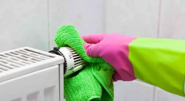Mano con guanto rosa e verde pulisce il termostato del termosifone con un panno verde. Guida su come pulire i termosifoni.